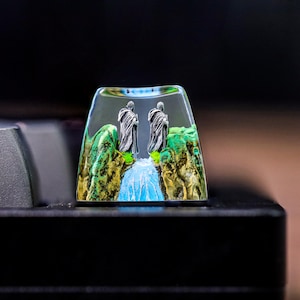 Puede incluir: Un keycap de resina transparente con una escena en miniatura de dos figuras de pie en un puente sobre una cascada. Las figuras llevan túnicas oscuras y el fondo es un paisaje verde y marrón.