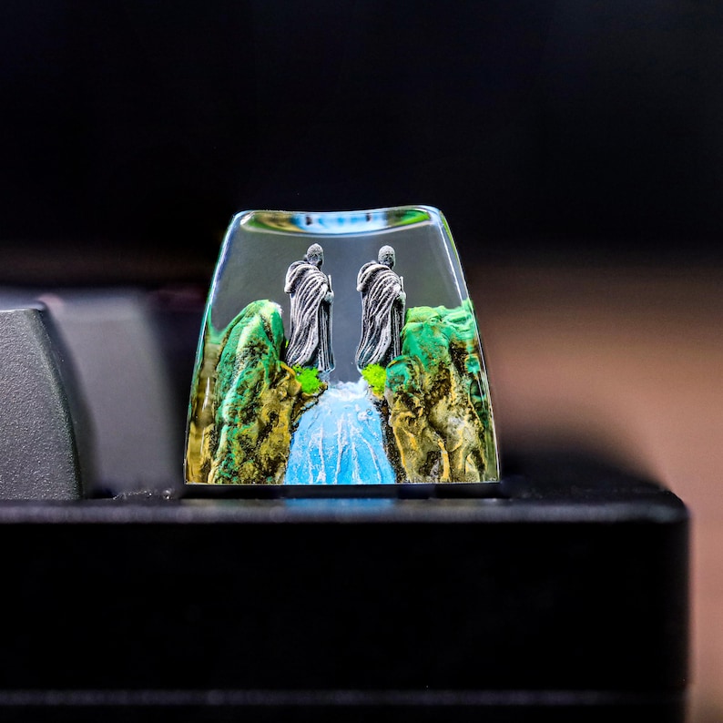 May include: A clear resin keycap with a miniature scene of two figures standing on a bridge over a waterfall. The figures are wearing dark robes and the background is a green and brown landscape.