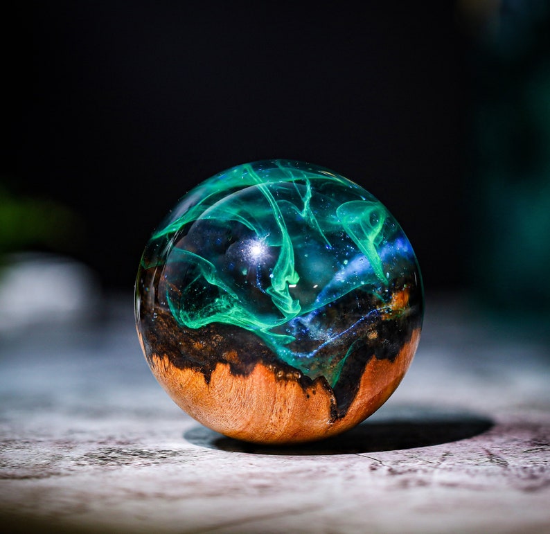 May include: A round, wooden sphere with a clear resin coating. The resin contains swirls of green and blue, resembling a galaxy. The sphere is approximately 2 inches in diameter.