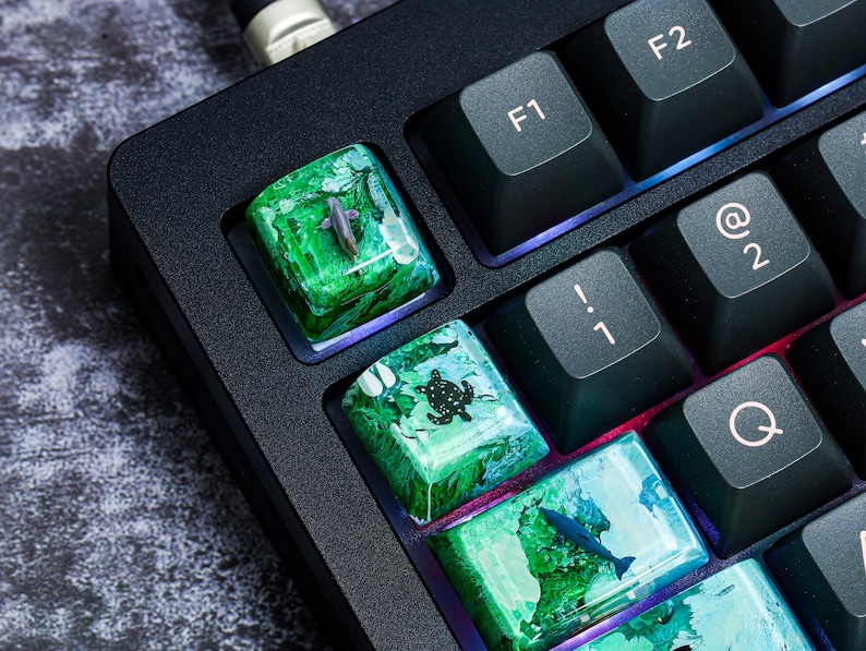 May include: A close-up of a black mechanical keyboard with custom keycaps featuring ocean-themed designs. The keycaps are made of resin and have a translucent, green and blue color scheme. The designs include a turtle, a fish, and a dolphin.