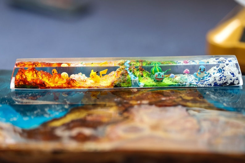 May include: A clear rectangular resin block with a layered landscape scene. The scene includes a yellow and orange mountain range, a green forest with a green and brown creature, and a blue ocean with a blue and white creature. The resin block is on a wooden surface.