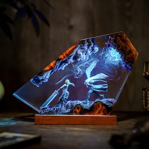 May include: A resin art piece featuring two figures in a battle scene, set within a clear, angled block. The artwork has a wooden base and incorporates blue and orange hues, with a glowing effect. The figures are in a dynamic pose, suggesting action.