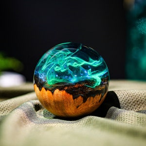 May include: A round, wooden orb with a clear resin top filled with swirling green and blue resin. The resin has a translucent, almost smoky appearance.