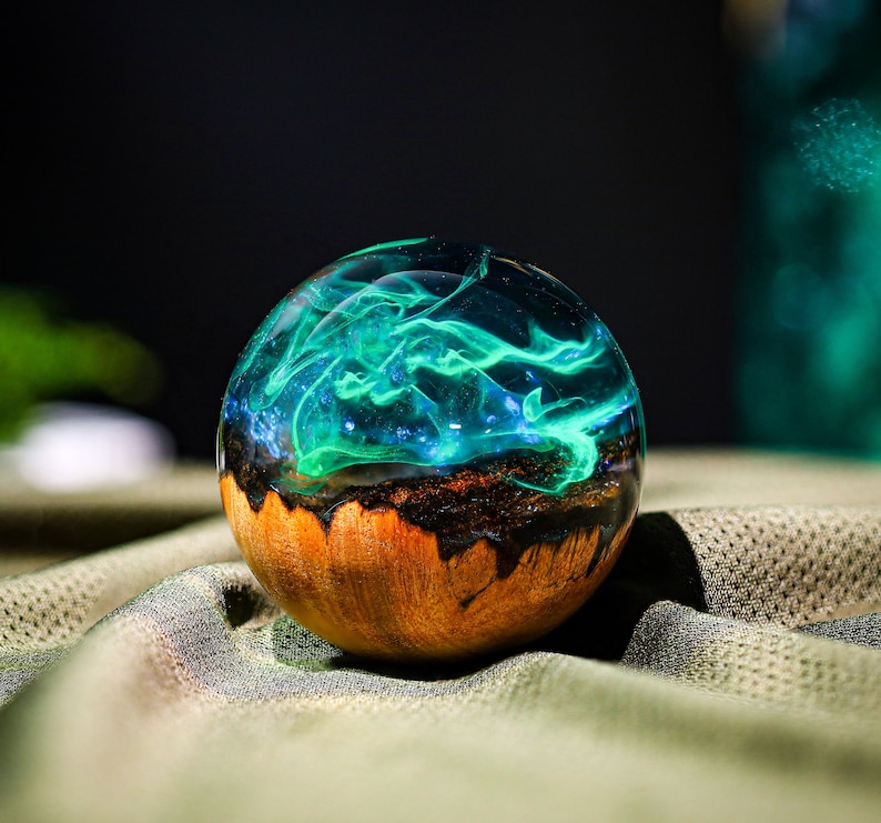 May include: A round, wooden orb with a clear resin top filled with swirling green and blue resin. The resin has a translucent, almost smoky appearance.