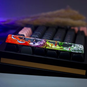 May include: A colorful resin keyboard wrist rest with a landscape scene featuring miniature figures. The wrist rest is made of clear resin with layers of orange, purple, green, and white. The scene includes a small waterfall, trees, and a path. The miniature figures are of people walking along the path.