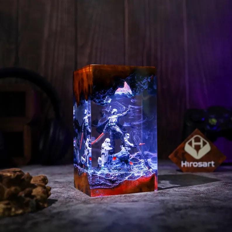 May include: A rectangular resin lamp featuring a dark wood base and top, encasing a miniature scene with a central figure holding a red lightsaber surrounded by white-armored soldiers. The interior glows with blue light, creating a nebula effect.