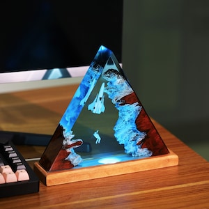 May include: A triangular resin sculpture with a blue and white abstract design, featuring a white airplane and a small figure, set on a wooden base.