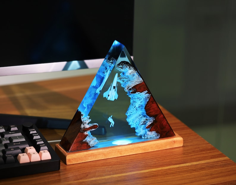 May include: A triangular resin sculpture with a blue and white abstract design, featuring a white airplane and a small figure, set on a wooden base.