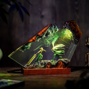 May include: A detailed resin sculpture featuring two figures in a battle scene, set within a clear, angled block. The figures are green, with one wielding a large sword. The base is a dark wood, and the scene is illuminated with green light.