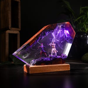 May include: A decorative resin sculpture featuring two figures with lightsabers in a dynamic pose, set against a swirling purple background. The sculpture is encased in a clear, angled resin block with a wooden base.