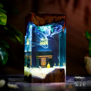 May include: A rectangular resin and wood lamp featuring a miniature black Kaaba model inside. The clear resin section contains swirling blue and purple smoke-like patterns and gold Arabic calligraphy. The base and top are made of dark wood.