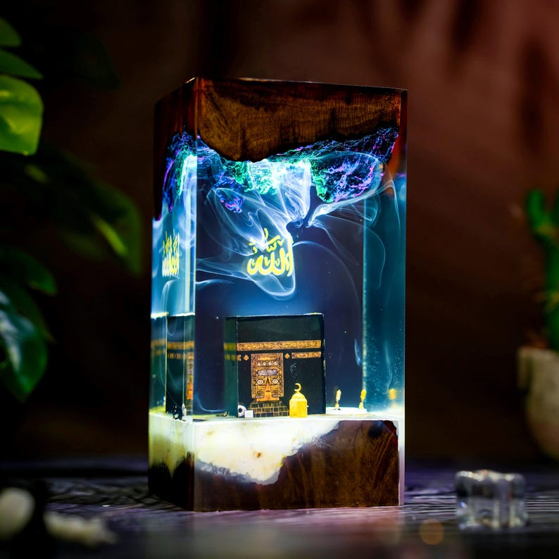 May include: A rectangular resin and wood lamp featuring a miniature black Kaaba model inside. The clear resin section contains swirling blue and purple smoke-like patterns and gold Arabic calligraphy. The base and top are made of dark wood.