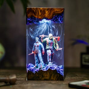 May include: A rectangular resin art piece featuring two horror movie figures, one with a mask and axe, the other with a knife. The figures are set against a smoky blue and white background, with a wooden base and top.