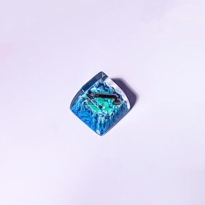 May include: A blue and green resin keycap with a silver keycap stem and a small silver keycap switch. The keycap is shaped like a square with rounded corners.