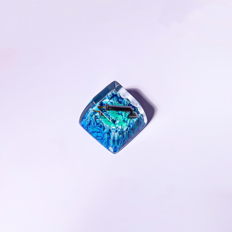 May include: A blue and green resin keycap with a silver keycap stem and a small silver keycap switch. The keycap is shaped like a square with rounded corners.