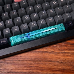 May include: A teal and blue keyboard keycap with a sword design. The keycap is made of resin and has a textured surface.