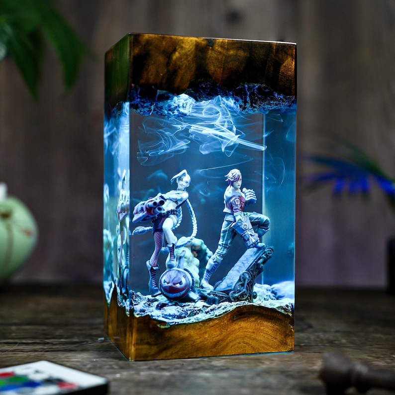May include: A rectangular resin lamp with a wooden base and top. Inside, two figurines are set against a blue, smoky background. The lamp features a unique design, combining wood and resin for a decorative lighting piece.