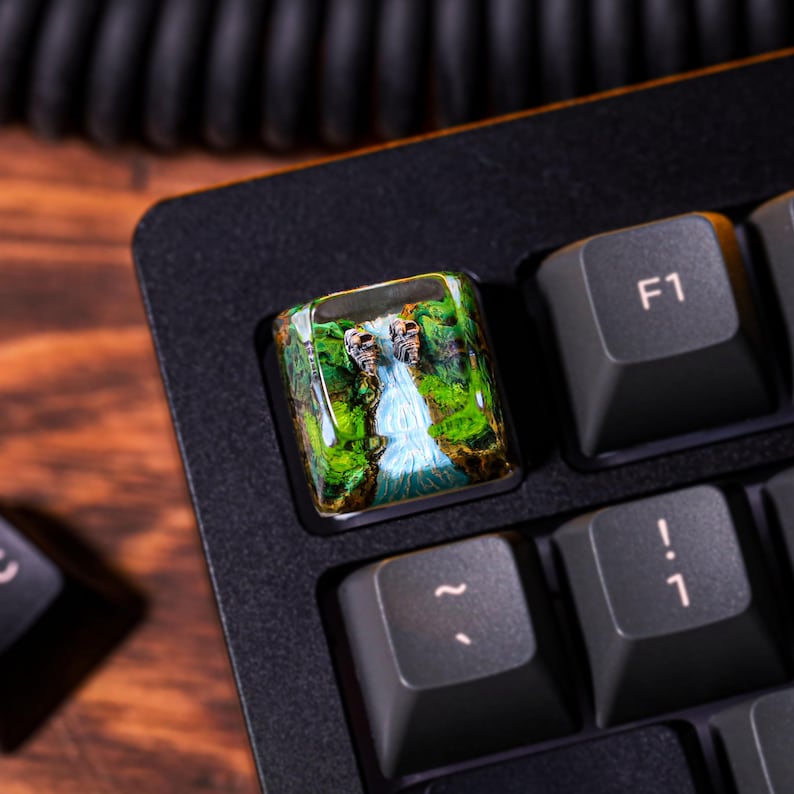 May include: A custom keyboard keycap with a green and blue landscape scene featuring a waterfall and two small figures. The keycap is made of resin and has a glossy finish.