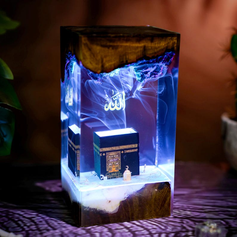 May include: A rectangular resin lamp featuring a dark wood top and base with a clear center section. Inside the resin is a miniature black Kaaba model, a small gold-colored lantern, and a glowing blue light effect with the Arabic word Allah.