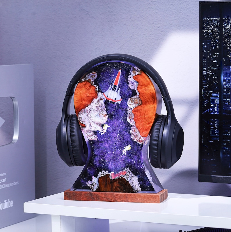May include: A black headphone stand with a galaxy-themed resin base. The base features a spaceship, stars, and a purple and blue nebula. The stand is made of wood and has a natural finish.