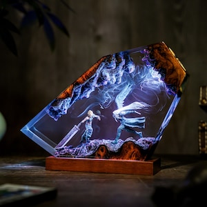 May include: A resin art piece featuring a battle scene with two figures, one wielding a large sword. The artwork is encased in a clear, angled block with a wooden base. The scene is illuminated with blue and white light.