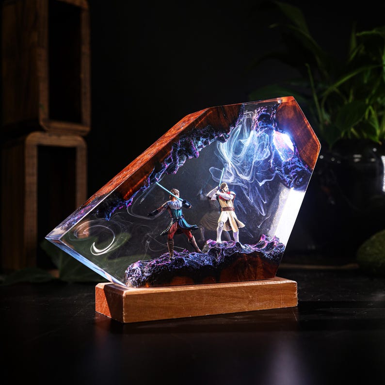 May include: A unique resin sculpture featuring two figures with glowing lightsabers in a dynamic battle pose. The sculpture is encased in a clear, angled resin block with a wooden base, showcasing a detailed, colorful scene.