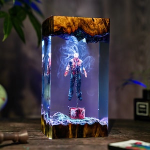 May include: A rectangular resin sculpture featuring a figure suspended by a chain. The figure is dressed in blue jeans and a blood-splattered shirt. The sculpture has a wooden base and top, with blue and white swirling accents.