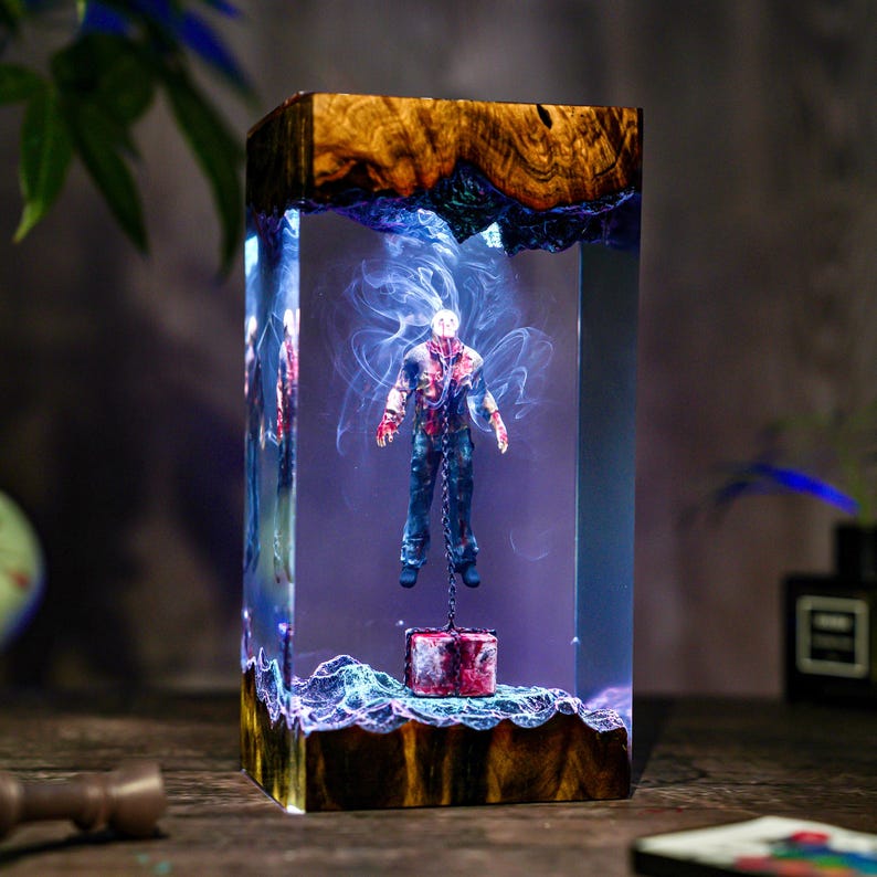 May include: A rectangular resin sculpture featuring a figure suspended by a chain. The figure is dressed in blue jeans and a blood-splattered shirt. The sculpture has a wooden base and top, with blue and white swirling accents.