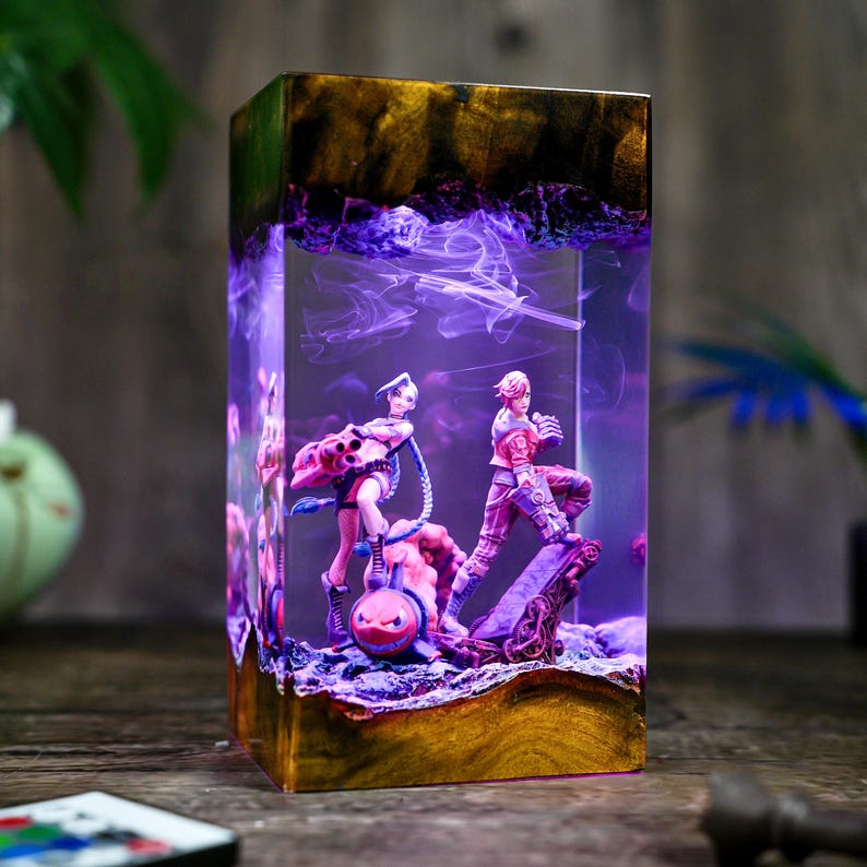 May include: A rectangular resin art piece featuring two figurines inside. The resin is clear with a purple, smoky effect and a wooden base. The figurines are of a woman and a man in action poses, with pink and red clothing.