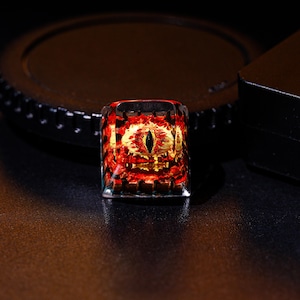 May include: A red and gold keycap with a black eye design. The keycap is made of clear resin and has a glossy finish.