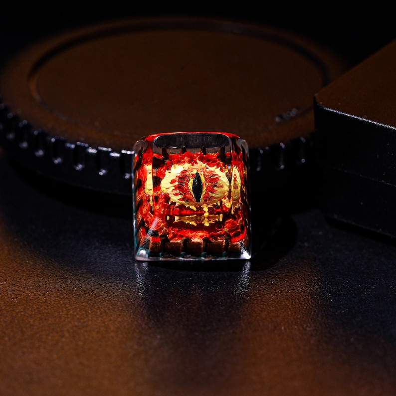 May include: A red and gold keycap with a black eye design. The keycap is made of clear resin and has a glossy finish.