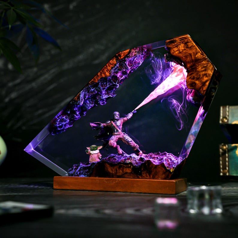 May include: A decorative lamp featuring a resin and wood frame with a purple and pink illuminated interior. Inside, a miniature armored figure in a dynamic pose projects a light beam, accompanied by a small green creature on a rocky base.