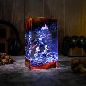 May include: A rectangular resin lamp featuring a dark wood top and base with a clear center section. Inside the resin, a black-clad figure stands on a rocky outcrop surrounded by white-armored figures, illuminated by a bright blue internal light.