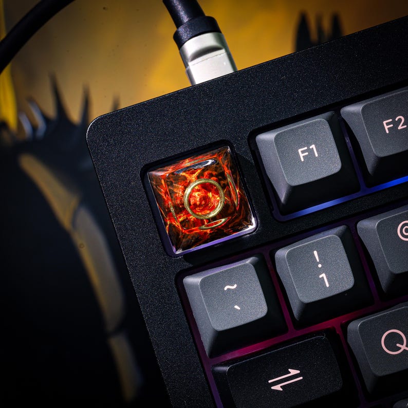 May include: A black mechanical keyboard with a custom keycap. The keycap is a square with a clear, red, and orange resin design. The keycap is labeled with the letter "F1".