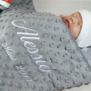 May include: A gray and white baby blanket with the name "Alessio" embroidered on it. The blanket is made of a soft, textured fabric and has a star pattern. A baby is sleeping on the blanket wearing a white hat with the words "Baby Boy" embroidered on it.
