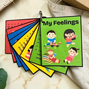 May include: A fan-style educational card set titled My Feelings, featuring colourful square cards bound by a metal ring. The top green card displays illustrations of children playing, reading, and relaxing, with black text on a bright green background.