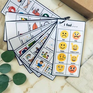 May include: A fan-style set of white communication cards bound by a metal ring. The top card features the text 'I feel...' and a grid of eight yellow emojis labelled with emotions like sad, sick, happy, ready to learn, nervous, excited, overwhelmed, and angry.