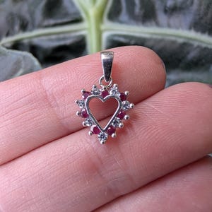 May include: A silver pendant in the shape of a heart, adorned with small red and clear gemstones. The heart-shaped pendant has a loop at the top for a chain. The pendant is held between fingers against a blurred green background.