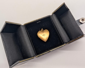 May include: A gold heart-shaped pendant with a small diamond accent, displayed in an open black jewelry box. The pendant is suspended from a delicate chain. The box is black with gold trim and a snap closure.