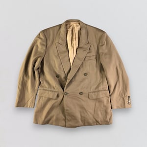 Puede incluir: Un blazer cruzado de color beige con solapa de muesca, dos bolsillos delanteros y cierres de botón. El blazer tiene un forro de color claro y botones en las mangas. Esta chaqueta clásica es adecuada para vestimenta formal o de negocios.