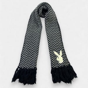 May include: A black and white knit scarf featuring a repeating herringbone pattern throughout the fabric. The scarf includes a light-colored embroidered rabbit logo on one end and is finished with black fringe trim at both ends of the garment.