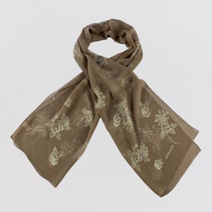 May include: A sheer brown rectangular scarf featuring a delicate, light-colored floral pattern of daisies and stems scattered across the fabric. The brand name ANNE KLEIN is printed in a small, subtle font near the bottom right corner of the scarf.