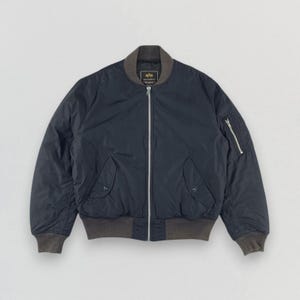 May include: Navy blue bomber jacket with a grey collar, cuffs, and hem. The jacket features a silver zip, two front pockets with snap closures, and a zipped pocket on the left sleeve. The jacket has a classic, streamlined design.