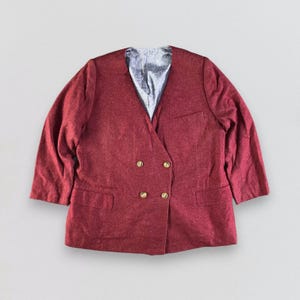 Puede incluir: Una blazer cruzada de color granate con botones dorados. La blazer tiene solapas con muescas y dos bolsillos delanteros. El forro interior es de tela plateada texturizada. Las mangas son de tres cuartos.