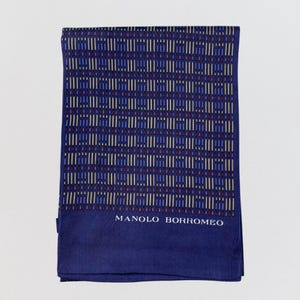 May include: A navy blue scarf with a geometric pattern of vertical lines in white, red, and light blue. The scarf has the text "MANOLO BORROMEO" in white at the bottom. The scarf is folded and displayed on a white background.
