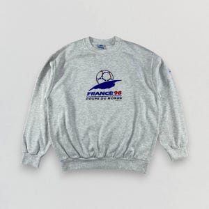 May include: A light gray sweatshirt featuring a graphic for the 1998 FIFA World Cup in France. The design includes a soccer ball, a blue stylized bird, and the text "FRANCE 98 COUPE DU MONDE" in blue and red.