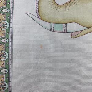 May include: A light gray fabric scarf featuring a partial illustration of a yellow elephant with a teal and white tusk. The border displays a repeating pattern of green, gold, and white floral motifs. A small, faint orange spot is visible on the fabric.