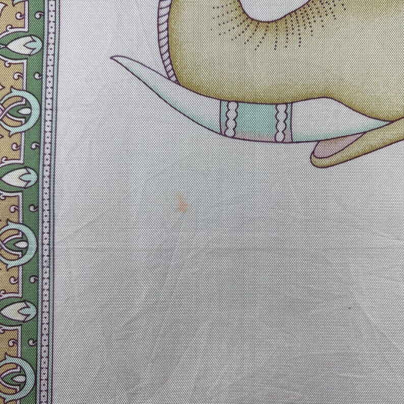 May include: A light gray fabric scarf featuring a partial illustration of a yellow elephant with a teal and white tusk. The border displays a repeating pattern of green, gold, and white floral motifs. A small, faint orange spot is visible on the fabric.