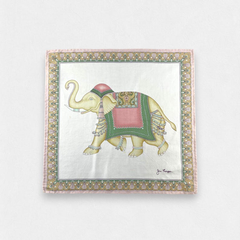 May include: A square silk scarf featuring a central illustration of a cream-colored elephant adorned with a pink and green decorative blanket, headpiece, and tassels. The scarf has a wide, ornate border in shades of gold, pink, and green.