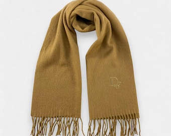 May include: A tan scarf with a fringed edge. The scarf is folded and draped, with a subtle embroidered logo near the bottom. The scarf is made of a soft material.